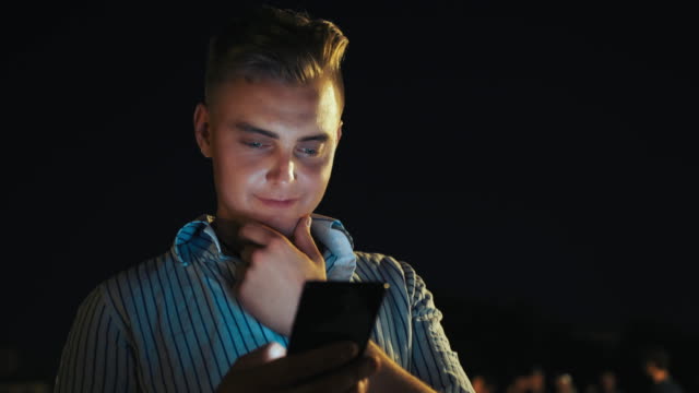 Close-up-portrait-of-young-businessman-using-his-modern-technologies-smartphone-at-night.-Handsome-man-browsing-internet-and-checking-mobile-apps-on-crowded-background.-Illuminated-face-from-phone-light-on-night-background.-Online-apps-and-network
