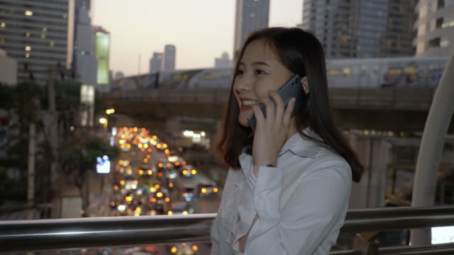 Junge-lächelnde-Geschäftsasiatische-Frau-mit-Smartphone-und-zu-Fuß-auf-der-modernen-Stadtstraße,-während-sie-mit-ihrem-Freund-auf-dem-Handy-in-der-Dämmerung-von-Bangkok-Thailand-sprechen.