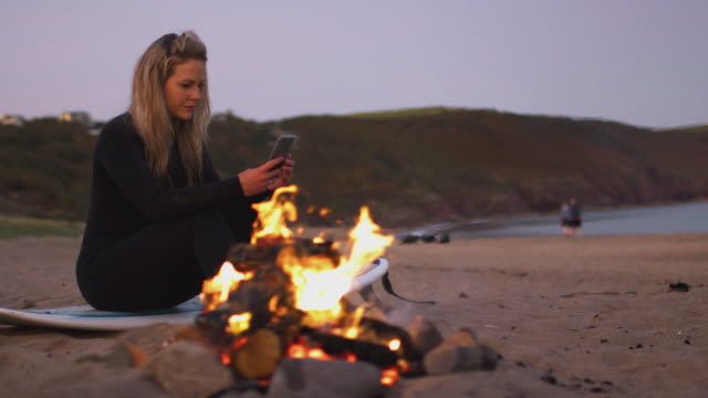 Frau-sitzt-auf-Surfbrett-von-Lagerfeuer-am-Strand-mit-Handy-als-Sonne-setzt-sich-hinter-ihr