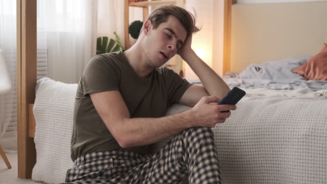 Man-yawning-and-text-messaging-on-mobile-phone-at-home