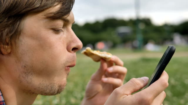 attraktiver-Mann-beißt-Scheibe-Pizza-und-verwendet-Smartphone-Nahaufnahme-im-Park-in-der-Natur