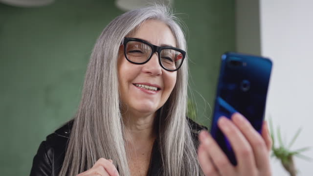 Close-view-of-charming-happy-senior-woman-in-glasses-with-long-gray-hair-which-having-video-call-using-blue-phone