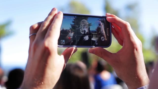Hand-fingers-hold-phone-making-video-of-wedding-ceremony-screen-close-up-slow-motion.-Smart-technologies-live-event-social-media-networks-guest-using-device-to-record-friends-couple-getting-married