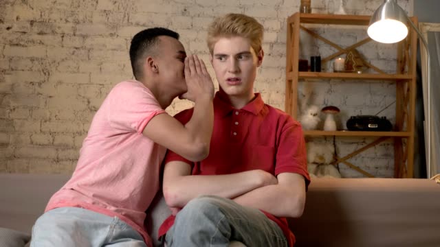 Zwei-junge-homosexuelle-Jungs-auf-der-Couch-sitzen,-sagt-afrikanischen-Mann-mit-kurzen-Haaren-die-absondern-zu-seinem-Partner,-die-Blondine-ist-schockiert-60fps