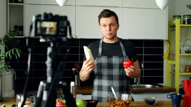 Cheerful-attractive-man-recording-video-food-vlog-about-healthy-cooking-on-digital-camera-in-the-kitchen-at-home.-Vlogging-and-social-media-concept