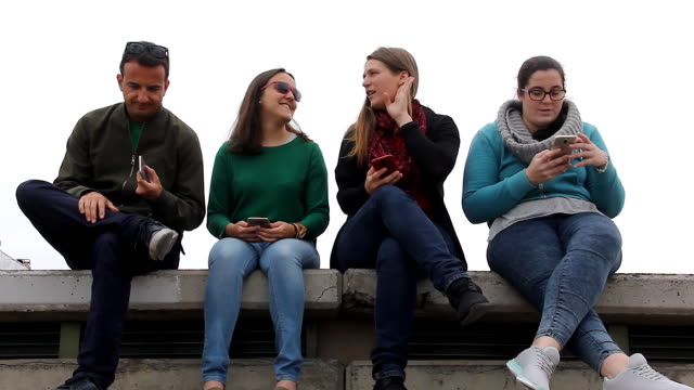 Drei-Frauen-und-ein-Mann-mit-Handys-sitzen-auf-Tribünen-an-bewölkten-Tag