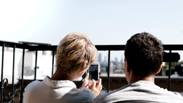 Happy-romantic-couple-sitting-at-a-small-sunny-balcony,-taking-smartphone-photos-of-amazing-Manhattan-view-in-New-York