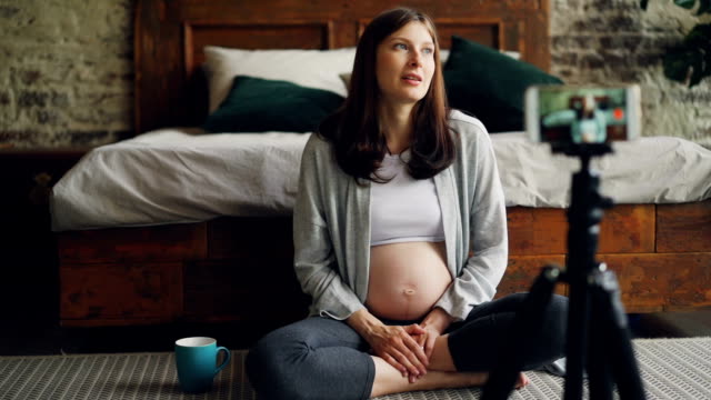 Bekannte-Blogger-ziemlich-schwangere-Frau-ist-für-Anhänger-mit-Blick-auf-Smartphone-Kamera-und-sprechen-auf-Boden-im-Schlafzimmer-sitzen-und-Lächeln-Videoaufnahme.