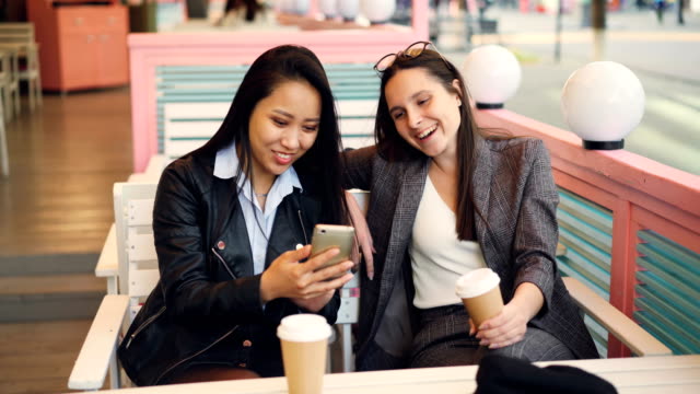 Muy-jovencitas-amigos-utilizan-moderno-smartphone-sentado-en-la-mesa-de-café-con-llevar-café.-Chicas-felizes-son-pantalla-táctil,-mirando-fotos-y-riendose.