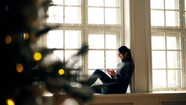 Happy-young-woman-is-using-smartphone-sitting-on-windowsill-on-Christmas-day-relaxing-at-home-and-enjoying-modern-technology.-People-and-gadgets-concept.