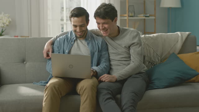 Adorable-Male-Gay-Couple-Spend-Time-at-Home.-They-Sit-on-a-Sofa-and-Use-the-Laptop.-They-Watch-Funny-Online-Videos.-Partner-Puts-His-Hand-Around-His-Lover.-Room-Has-Modern-Interior.