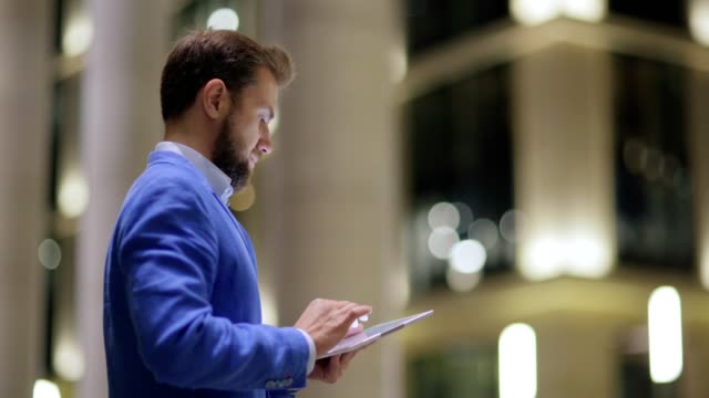 Seitenansicht-des-gut-aussehender-Mann-in-blauer-Jacke-mit-Tablet-Computer-im-Freien-im-Abend
