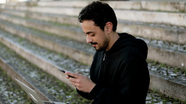 Joven-árabe-hombre-mensajes-de-texto-en-el-teléfono,-sosteniendo-portátil-en-las-rodillas