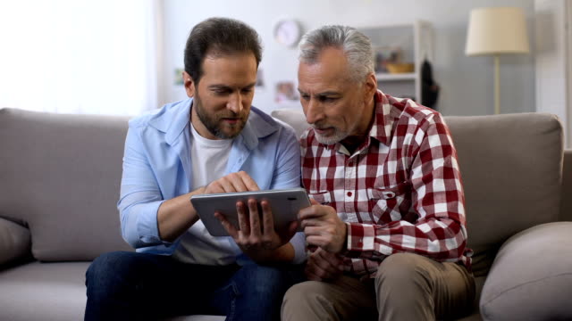 Retiree-father-and-middle-aged-son-scrolling-tab,-looking-and-smiling-to-camera