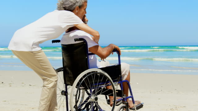 Vista-lateral-de-la-mujer-afroamericana-activa-que-abraza-a-un-hombre-mayor-con-discapacidades-en-la-playa-4k