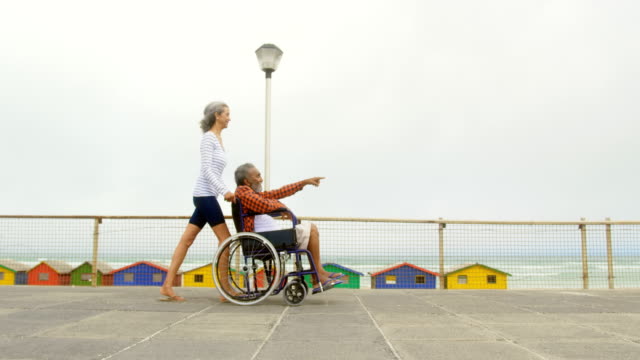 Seitenansicht-einer-aktiven-älteren-Afroamerikanerin,-die-behinderte-Mann-im-Rollstuhl-auf-Promenade-4k-schiebt