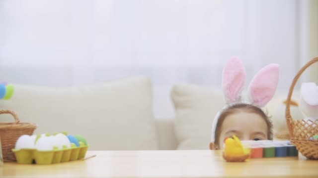 Little-playing-girl-with-bunny-ears-on-his-head-is-hiding-under-the-wooden-table,-full-of-Easter-decorations.-Girl-is-playing-showing-slolwly-her-charming-face.
