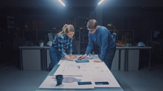 In-the-Dark-Industrial-Design-Engineering-Facility-Male-and-Female-Engineers-Talk-and-Work-on-a-Blueprints-Using-Conference-Table.-On-the-Desktop-Drawings,-Drafts-and-Electric-Engine-Components,-Parts