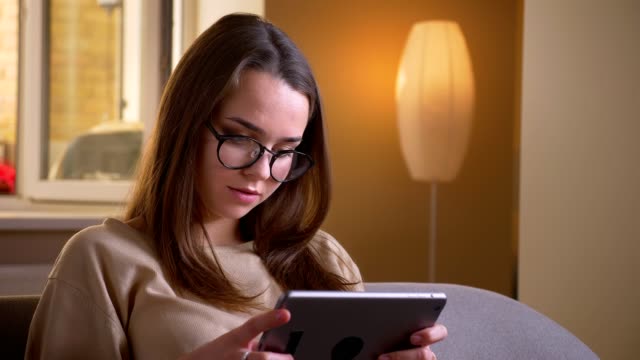 Closeup-portrait-of-young-attractive-caucasian-female-in-glasses-using-the-tablet-sitting-on-the-couch-indoors-in-a-cozy-apartment