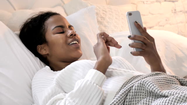 Mujer-africana-joven-que-usa-el-teléfono-inteligente-mientras-se-acuesta-en-la-cama