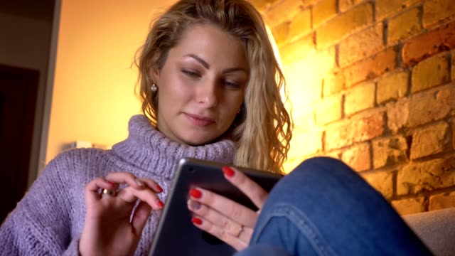 Closeup-front-shoot-of-adult-caucasian-blonde-female-texting-on-the-tablet-while-sitting-on-the-couch-indoors-in-a-cozy-apartment