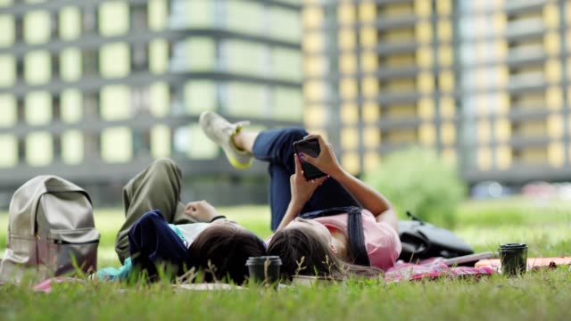 Zwei-Studentinnen-entspannen-sich-nach-dem-Unterricht-auf-dem-Rasen-auf-dem-Campus.-Mädchen-liegen-auf-Decke,-surfen-Smartphone-und-reden
