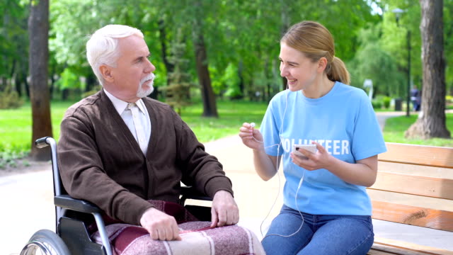 Happy-young-woman-giving-earphone-old-disabled-man,-supporting-hospital-patient