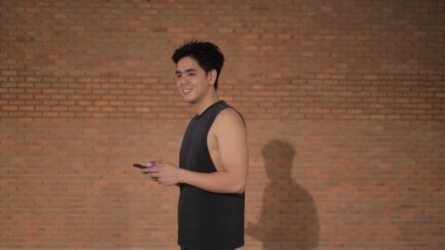 Young-happy-handsome-Asian-man-thinking-while-using-phone-against-brick-wall
