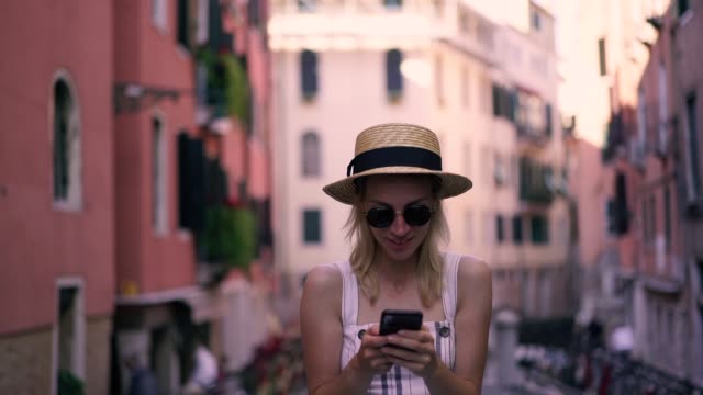Slow-motion-effect-of-cheerful-hipster-girl-in-stylish-sunglasses-typing-text-message-for-sending-to-friend-during-leisure-time-on-Venice-urban-setting