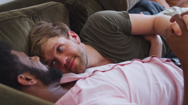 Loving-Male-Gay-Couple-Relaxing-Lying-On-Sofa-At-Home-Hugging-And-Kissingæ