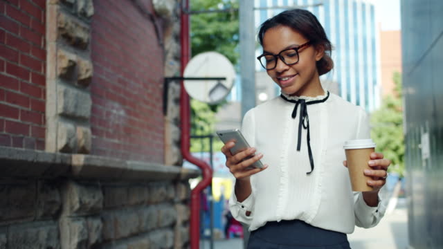 Sonriendo-mujer-afroamericana-usando-teléfono-inteligente-sosteniendo-café-caminando-al-aire-libre