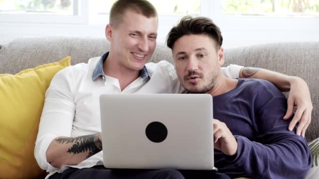 Gay-couple-relaxing-on-couch-using-laptop-computer.-Looking-at-screen.