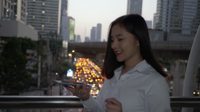 Junge-lächelnde-Geschäftsasiatische-Frau-mit-Smartphone-in-der-modernen-Innenstadt-in-der-Dämmerung-von-Bangkok-Thailand.-Sie-benutzt-ihren-Finger,-um-den-Bildschirm-zu-berühren.-Zum-Tragen-von-Hologrammen-entwerfen-Sie