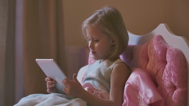 Portrait-of-pretty-blond-Caucasian-girl-lying-in-pink-bed-and-using-tablet.-Modern-child-watching-cartoons-before-going-to-bed.-Resting-at-home,-carefree-childhood.
