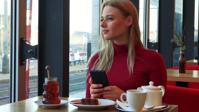 Eine-junge-attraktive-Frau-sitzt-an-einem-Tisch-mit-Essen-in-einem-Café-und-arbeitet-auf-einem-Smartphone-mit-einem-Lächeln,-schließlich-schaut-aus-dem-Fenster