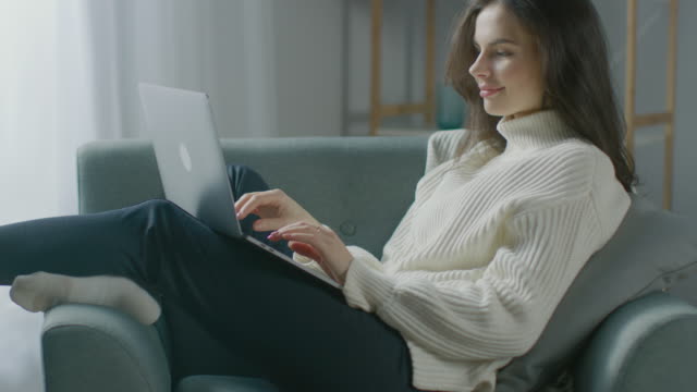 Hermosa-mujer-joven-trabaja-en-el-ordenador-portátil-mientras-está-sentada-en-la-silla.-Sensual-chica-usando-suéter-funciona-en-cuaderno;-Estudios,-Surfs-Internet,-Utiliza-las-redes-sociales-mientras-se-relaja-en-acogedor-apartamento