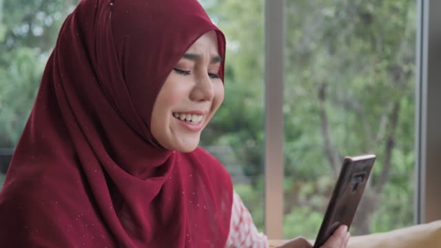 Glückliche-muslimische-Frau-mit-Smartphone-für-Videoanruf-zu-Hause.