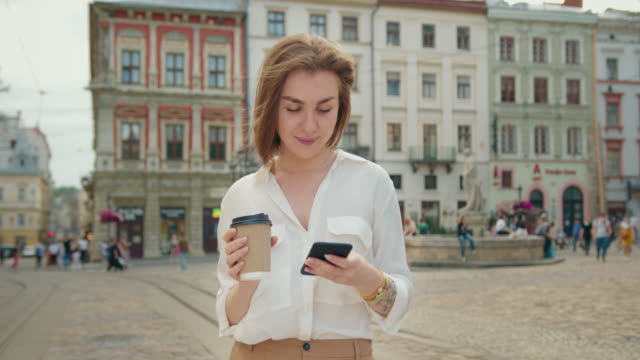 The-girl-is-texting-on-the-phone.-She-is-standing-and-smiling.-She's-holding-a-cup-of-coffee.-The-sun-is-shining-and-people-are-walking-in-the-background.-4K