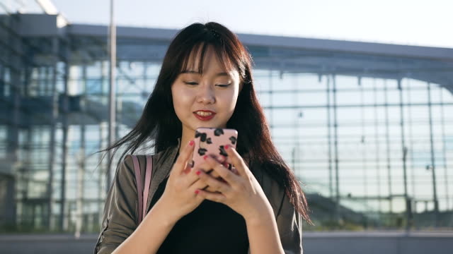 Nahansicht-der-jocund-glücklich-25-alter-asiatische-Dame-mit-langen-Haaren-und-stilvolle-Kleidung,-die-mit-ihrem-Smartphone-in-der-Nähe-von-modernen-Flughafengebäude