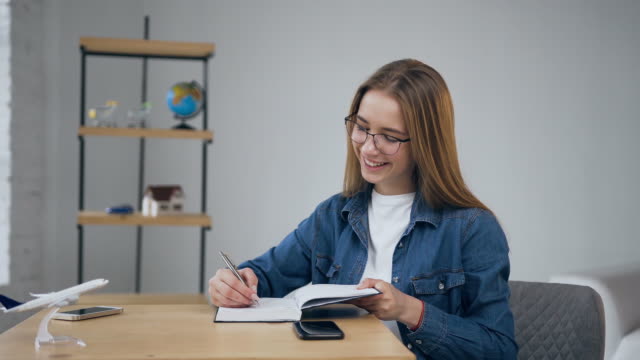 Cute-young-woman-putting-smart-phone-on-the-table-and-start-writing-in-the-notebook-at-home