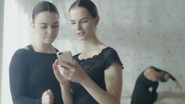Dos-Ballerinas-usando-smartphone-ordenador-mientras-tercer-ejercicios-en-la-barre-en-el-fondo.-Filmada-en-una-mañana-soleada-en-un-estudio-luminoso-y-moderno.-En-slow-motion.