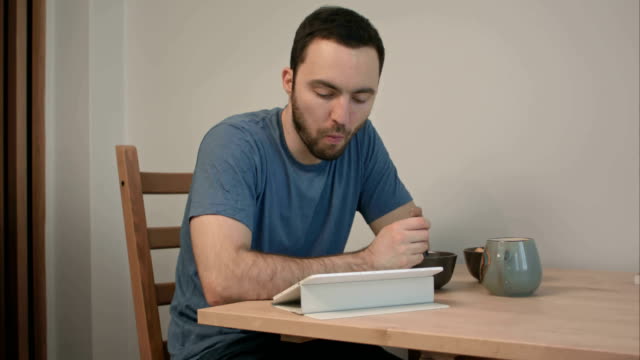 Junger-Mann-Essen-Frühstück-beim-Lesen-von-Nachrichten-auf-einem-Tabletcomputer