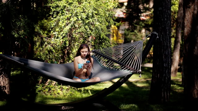 A-girl-in-a-blue-swimsuit-uses-her-phone-and-swings-in-a-hammock-in-nature.
