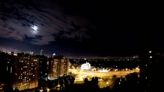 Moon-night-city-timelaps-in-moonlit-night-of-a-residential-area-near-the-high-speed-highway-with-fast-cars.-Clouds-passing-by-fuul-moon-at-night-city.-Nightclouds-fly-swiftly-across-the-sky