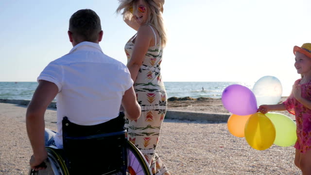 kid-with-Colorful-air-balloons-walks-near-mom-and-father-in-wheelchair,-invalid-with-wife-and-daughter-have-good-time