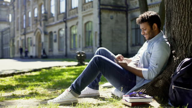 Multiracial-young-man-sitting-under-tree,-scrolling-mobile-phone-screen,-smiling