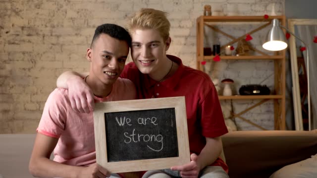 A-sad-international-gay-couple-is-sitting-on-the-couch-and-holding-a-sign.-We-are-strong.-Look-at-the-camera.-Home-comfort-on-the-background.-60-fps