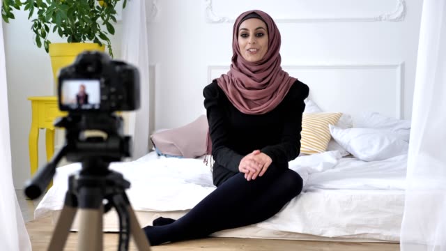 Young-beautiful-indian-girl-in-hijab-blogger-talking-on-camera,-gesturing,-white-room,-home-comfort-in-background.-50-fps