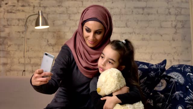 Junge-schöne-Mutter-Hijab-mit-kleinen-Mädchen-auf-der-Couch,-lächelnd,-Smartphone-nutzt,-macht-Selfie,-Kuscheln,-kleines-Mädchen-mit-Teddybär,-home-Komfort-im-Hintergrund-50-fps