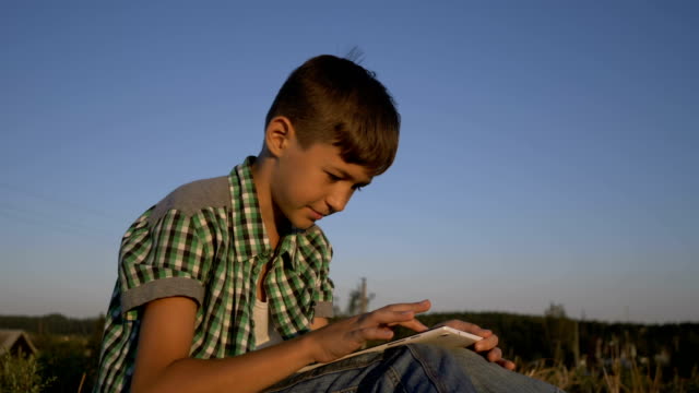 boy-use-tablet-sitting-in-field-at-sunset,-outdoors